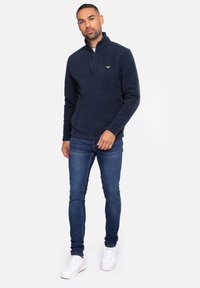Pullover in pile blu navy con cerniera a un quarto, caratterizzato da un piccolo logo giallo sul petto, abbinato a jeans in denim scuro e sneakers bianche.