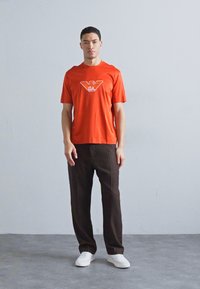 Raudona medvilninė marškinė su baltu logotipu, derinama su tamsiai rudos spalvos raukšlėtais kelnais ir baltais sportiniais bateliais, pastatyta prieš šviesiai pilką foną.