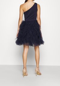 Tummansininen yksittäinen olkapäämekko, jossa on istuva pääntie ja röyhelöinen tyllihame. Yhdistetty beigen värisiin nilkkahihkaisiin korkokenkiin.