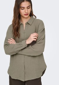 Olivengroen overhemd met knopen, gemaakt van een gestructureerde stof, met lange mouwen en een kraag. Heeft een afgeronde zoom en subtiele knoopdetails.