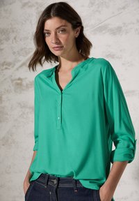 Blusa verde leggera con scollo a V, chiusura a bottoni e maniche a tre quarti. Tessuto morbido e vestibilità casual. Abbinata a jeans scuri.