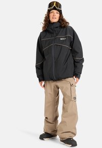 Juodas vandeniui atsparus švarkas su šoninėmis juostelėmis, užtrauktuku ir užtrauktukais užsegamomis kišenėmis, derinamas su smėlio spalvos cargo kelnėmis, turinčiomis šonines kišenes.