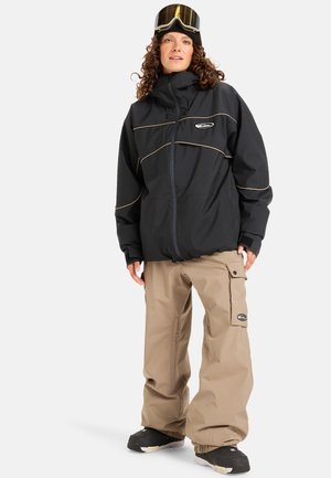 Giacca da neve nera con accenti beige, chiusura a zip e colletto alto, abbinata a pantaloni impermeabili beige e stivali da neve neri.