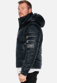 Giacca in pelle nera imbottita con cappuccio, dotata di tasche con zip sulle maniche e di una patch con logo sul braccio. Texture liscia e design aderente.