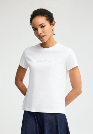 Frau mit kurzen lockigen Haaren, die ein weißes "Polo Club"-T-Shirt und einen dunkelblauen Rock trägt und mit den Händen hinter dem Rücken steht.