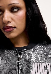 Szürke flitteres bomberdzseki cipzáras záródással, fekete gallérral és finom logórészlettel az elején. Simább, tükröződő felületű.