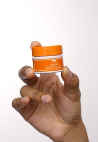 Pot orange de crème pour les yeux Brightening Eye Cream de Origins Ginzing avec des accents blancs, présentant une texture lisse et des détails sur l'étiquette, tenu dans une main.