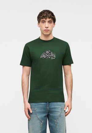 Muž oblečený v tmavozelenom tričku s grafikou osmičky a panorámou mesta, v kombinácii so svetlomodrými džínsami, stojaci na jednoliatom sivom pozadí.