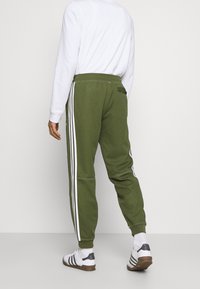 Man som bär olivgröna träningsbyxor med vita sidostripes, en vit långärmad tröja, vita strumpor och vita sneakers med svarta ränder.