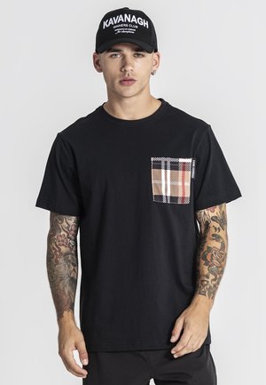 Jeune homme portant une casquette noire avec un texte blanc et un t-shirt noir avec une poche poitrine à motif à carreaux, affichant des avant-bras tatoués.