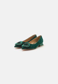 Ballerine in suede verde con punte affusolate, decorate con un fiore di pietre preziose sulla parte anteriore, dotate di tacco basso e quadrato con dettagli decorativi.