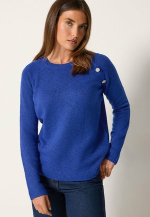Femme aux longs cheveux bruns portant un pull bleu avec des boutons argentés sur l'épaule et un jean bleu foncé, debout devant un fond uni.