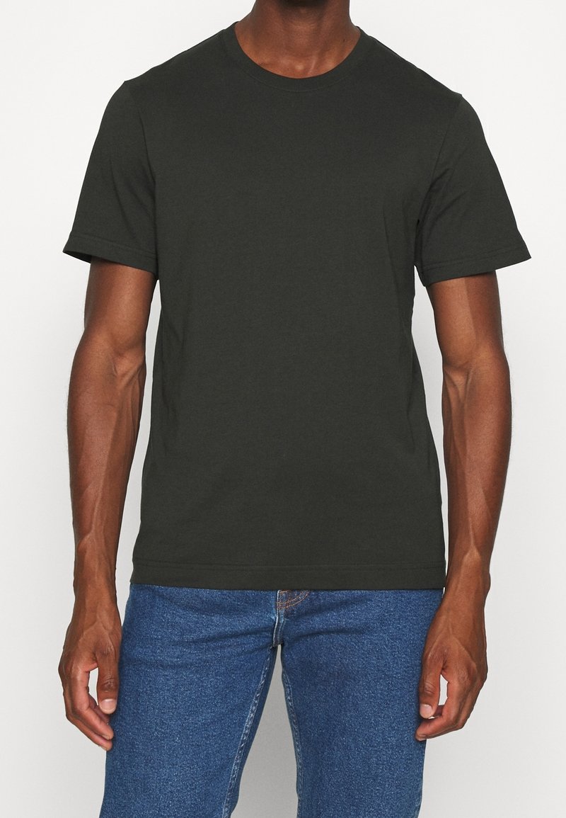 Homme portant un t-shirt noir uni à manches courtes et un jean bleu, debout, les bras détendus le long du corps.