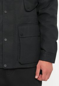 Black waterproof jacket with a zip and snap button closure, featuring a large side pocket and smooth, matte texture. Hand resting on pocket.
