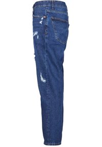 Jean en denim bleu avec une coupe décontractée, vue de côté, avec des empiècements usés et une poche arrière sur fond blanc.