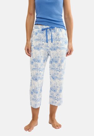 Vrouw draagt witte korte broek met blauw bloemmotief en blauwe trekkoord, gecombineerd met een mouwloos blauw rib topje, staat op blote voeten.