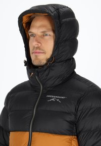 Svart puffjacka med en orange nederpanel, som har huva, dragkedja framtill och logotyper. Texturen är slät med sömnadsmönster.