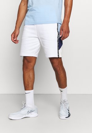 Białe szorty z niebieskim bocznym panelem i czarnym paskiem z logo. Noszone z jasnoniebieskimi tenisówkami i białymi skarpetkami. Swobodny krój i faktura.