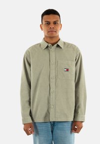 Green corduroy button-up shirt with a chest pocket, featuring a small multicolored logo. Paired with light blue jeans.