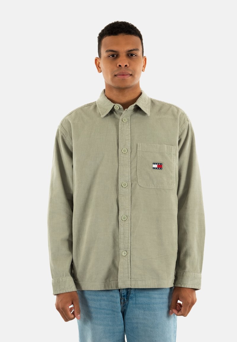Green corduroy button-up shirt with a chest pocket, featuring a small multicolored logo. Paired with light blue jeans.