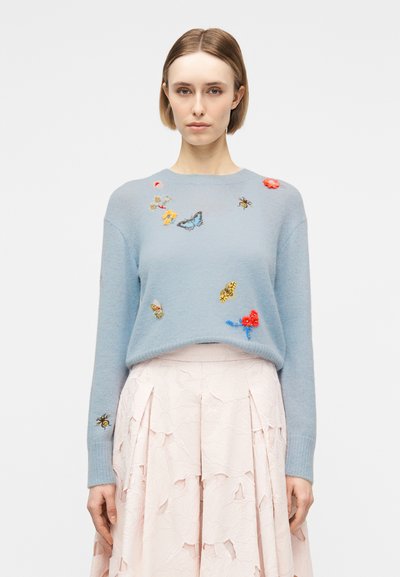 Vrouw met kort haar, gekleed in een lichtblauwe trui met geborduurde insecten en bloemen, gecombineerd met een lichtroze textuurrijke rok.