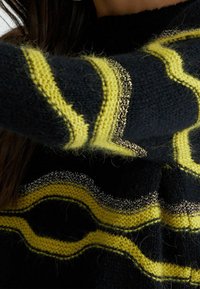 Femme portant un pull noir duveteux avec des rayures zigzag jaunes et argentées métalliques, couvrant partiellement son cou et ses bras.