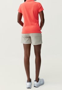 Korallfärgad pikétröja, beige shorts, vita och gråa träningsskor. Bilden visar baksidan med korta ärmar och enkel design utan synliga mönster.