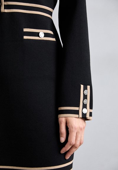 Robe noire avec des rayures horizontales crème, une poche sur la poitrine et trois boutons argentés au poignet, le tout avec une texture lisse.