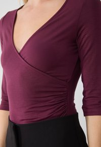 Burgundy long-sleeve top with a V-neck and ruched side detailing, made from soft, stretchy fabric, paired with black high-waisted pants.