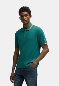 Polo in teal con un accento arancione a contrasto sul colletto. Maniche corte, texture liscia e dettaglio del logo sulla manica sinistra.