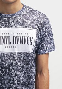 Tričko s krátkými rukávy v černobílém květinovém vzoru, s bílým obdélníkovým grafickým prvkem uprostřed, ve kterém je černý text.