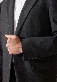 Blazer gris oscuro con textura sutil, solapas de pico y cierre de un solo botón. Presenta un bolsillo frontal y cuatro botones en la muñeca.