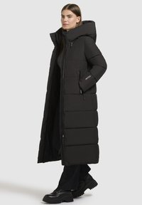 Moteris, vilkinti ilgu juodu pūkuotu paltu su aukštu apykakle ir kapišonu, juodomis kelnėmis ir juodais masyviais platforminiais batais, stovinti šonu.