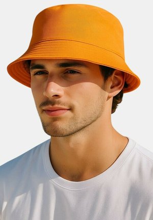Jonge man met lichte gezichtshaar, die een oranje vissershoed en een wit T-shirt met ronde hals draagt tegen een effen achtergrond.