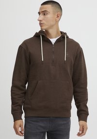Brown zip-up hoodie with a front pocket, drawstring hood, and subtle logo detail. Made of soft, textured fabric. Pairing with gray jeans.