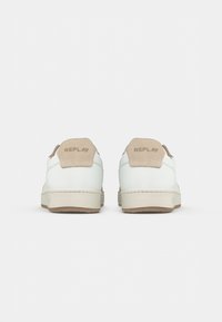 Une paire de baskets blanches avec des onglets de talon beige embossés du nom de la marque "Replay", présentée de dos sur un fond blanc.