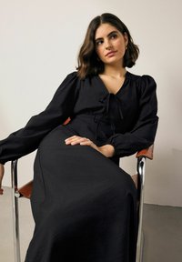 Černé šaty s dlouhým rukávem mají hluboký výstřih se zavazováním a splývavý siluetu, na neutrálním pozadí.