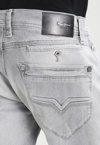Detailní záběr na zadní kapsu světle šedých džínsů s koženou nášivkou Pepe Jeans London a kovovými nýty v rozích kapsy.