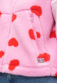 Φλις ζακέτα σε ροζ χρώμα με κόκκινες καρδιές. Διαθέτει ροζ φερμουάρ και ένα μικρό λογότυπο στο στρίφωμα. Μαλακό, υφασμάτινο ύφασμα.
