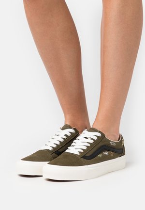 Persona che indossa sneakers in suede verde oliva con lacci e suole bianche, dotate di piccoli motivi ricamati sui pannelli laterali.