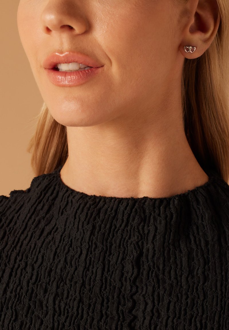 Boucles d'oreilles en argent en forme de cœurs entrelacés, présentées sur un tissu noir texturé. Fini lisse avec un éclat poli.