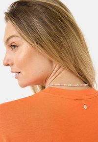Orange bomullströja med rund halsringning och en dekorativ metallaccent på baksidan. Har en flerfärgad pärlhalsband.