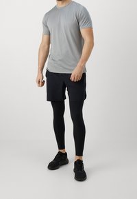 Camisa deportiva de hombre de color gris claro y de manga corta, combinada con pantalones cortos negros y mallas de compresión negras. Zapatillas deportivas completan el conjunto.