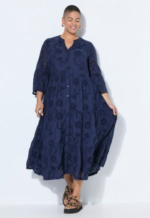 Kvinde iført en mørkeblå, langærmet kjole med knapper og broderede blomster, iført sandaler med leopardmønster, stående op ad en hvid væg.