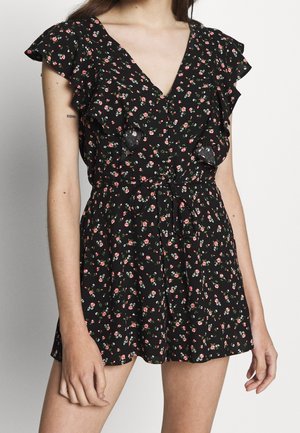 Woman wearing a black floral romper with ruffled sleeves, V-neckline, and a waist tie, standing against a plain light background.