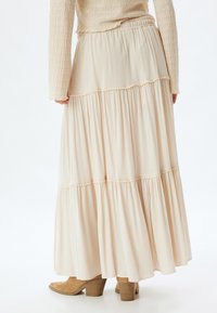 Jupe maxi tiered couleur crème avec un tissu fluide, une texture subtile et une taille froncée. Associée à des bottes à talons marron clair.
