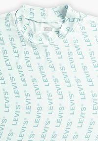 Světle modré tričko s vysokým výstřihem, které zdobí opakovaný nápis "LEVI'S" tučným mátově zeleným písmem. Hladká látka s jemným leskem.