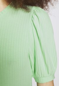 Maglietta verde chiaro a coste con maniche puff, scollatura rotonda, tessuto testurizzato e polsini elastici. Ingrandimento del tessuto e dei dettagli.