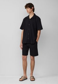 Schwarzes kurzärmliges Hemd mit Kragen und passende Shorts aus strukturiertem Stoff, kombiniert mit braunen Slippers mit Schnalle.