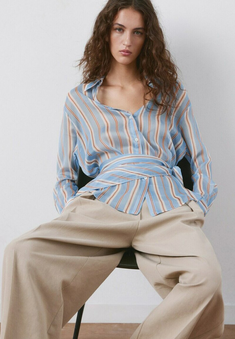 Jeune femme assise portant un pantalon beige ample et une chemise à rayures bleu clair boutonnée avec une taille nouée, sur un fond uni.